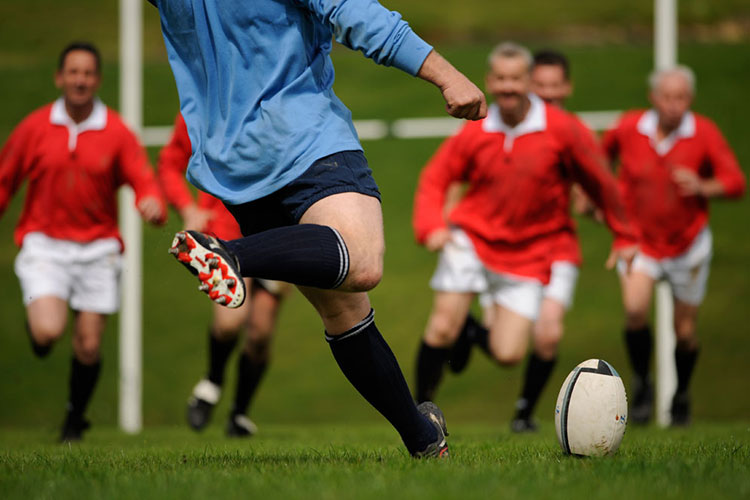 A rugby player in a blue polo gets ready to kick the ball with opponents in red polos running towards him