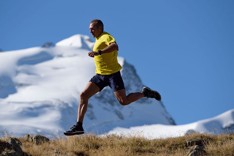 An older man in a yellow shirt and blue shorts sprints across a trail