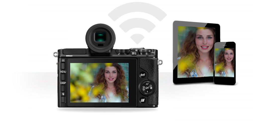 Sharing wirelessly with the Nikon 1 V2 and WU-1b Image of the Nikon 1 V2 and WU-1b along with a table and smartphone, and the wireless transmission symbol showing Wi-Fi sharing
