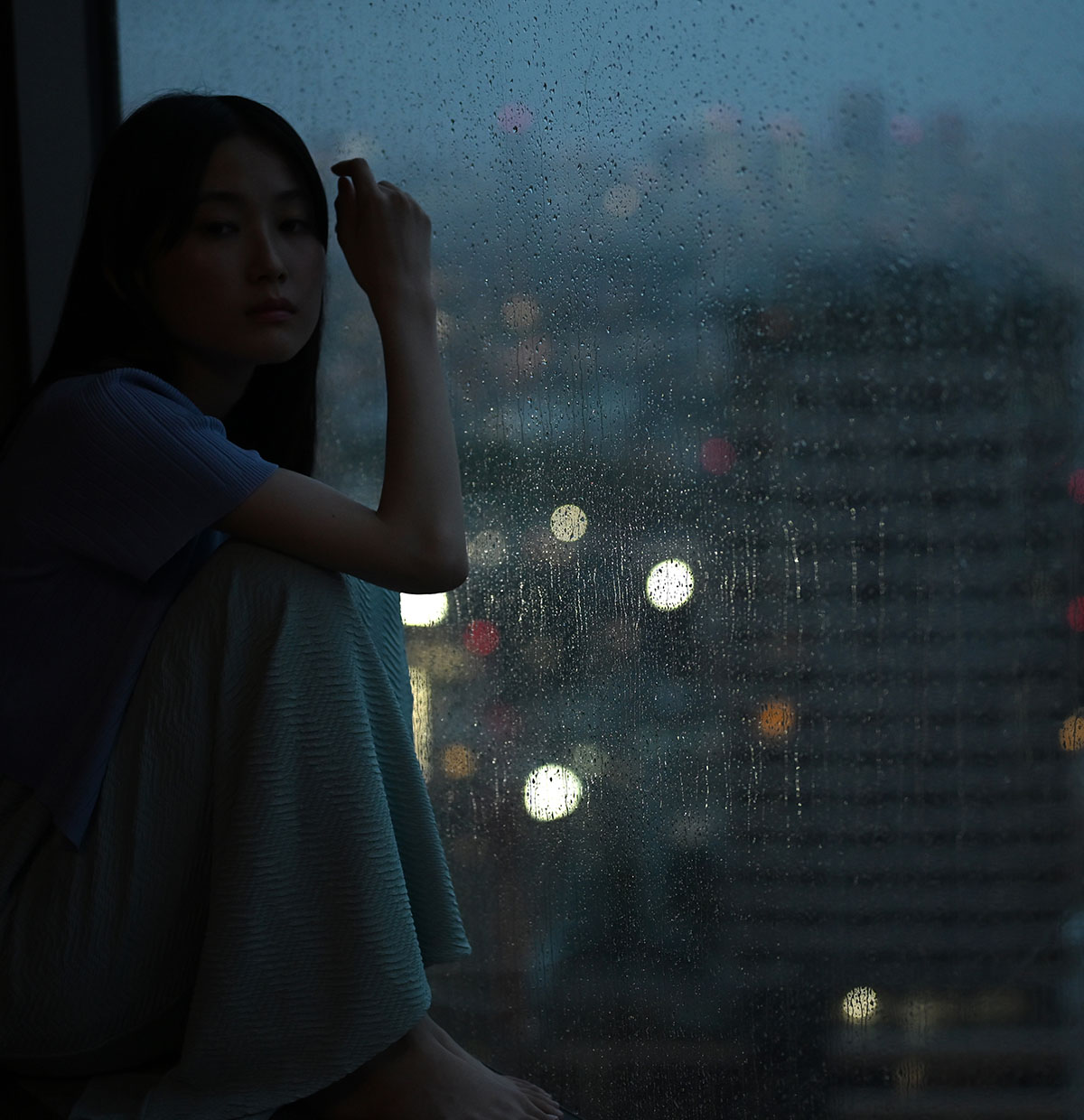 Photo of a female model in low light in front of a window with rain drops on it, taken with the Z f camera