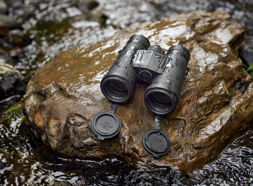 A pair of Nikon Monarch M5 binoculars on a rock, with splashes of water on the rock and binoculars