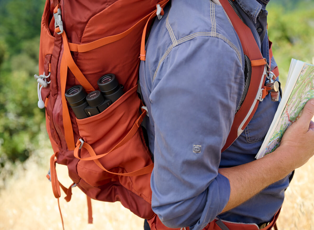 Photo of a person's torso wearing a backpack, holding a map with a pair of Monarch M7 binoculars in the bag