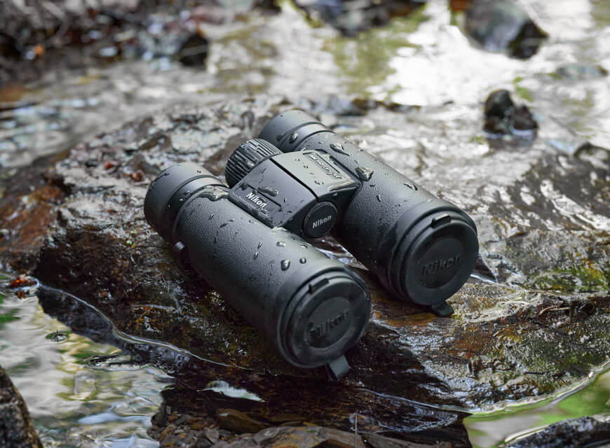 A pair of Nikon Monarch M7 binoculars on a rock, with splashes of water on the rock and binoculars